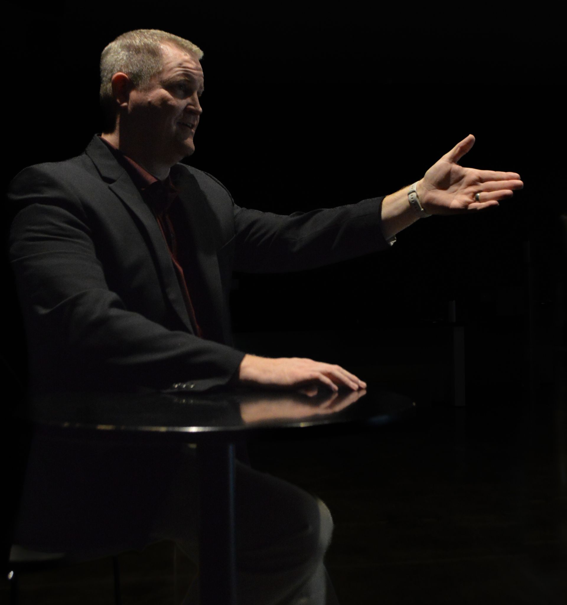 Man in a suit gestures with his right hand, sitting at a dark table.