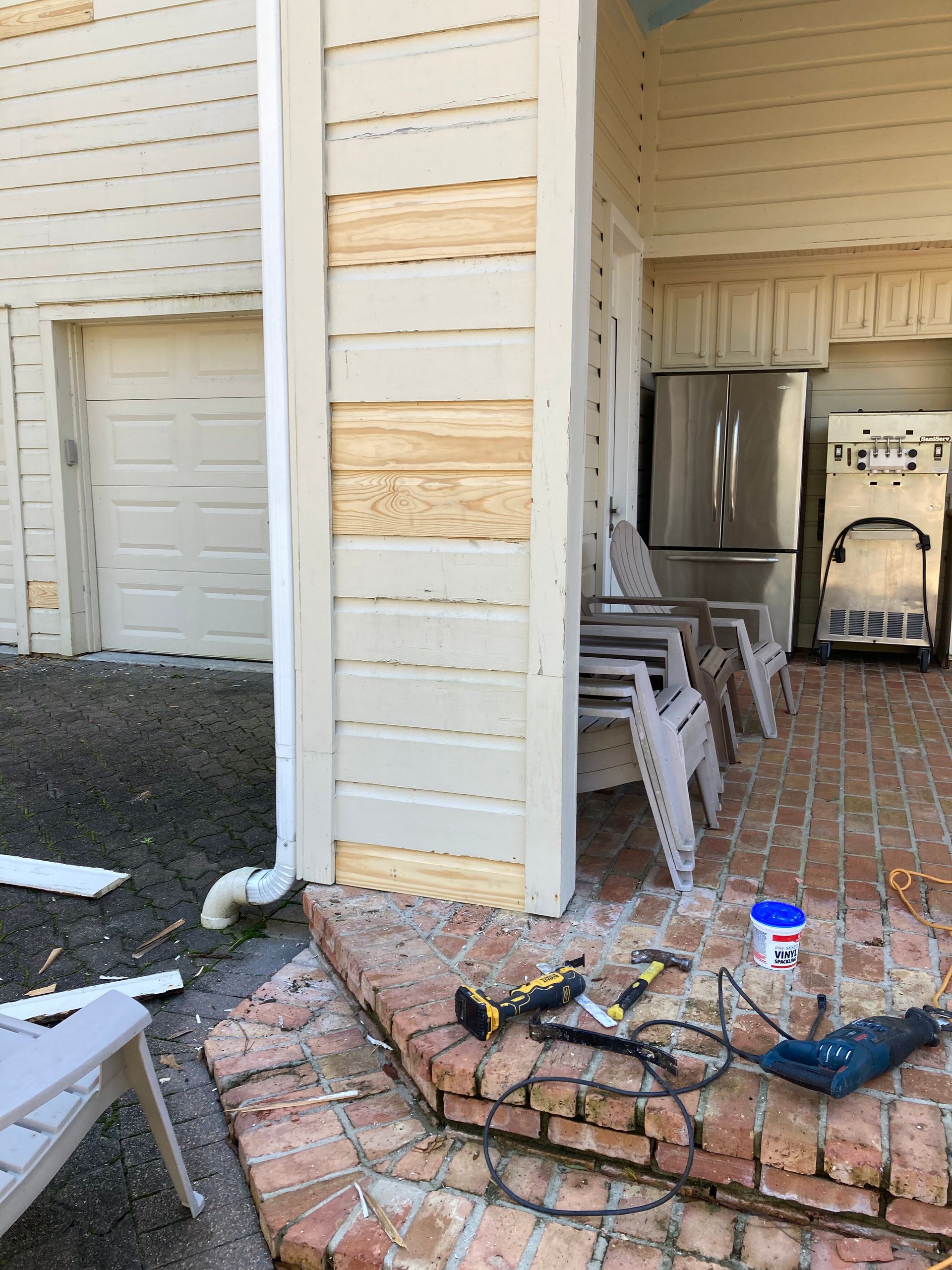 Damaged porch with broken siding, tools on the steps, and stacked chairs beside a garage door