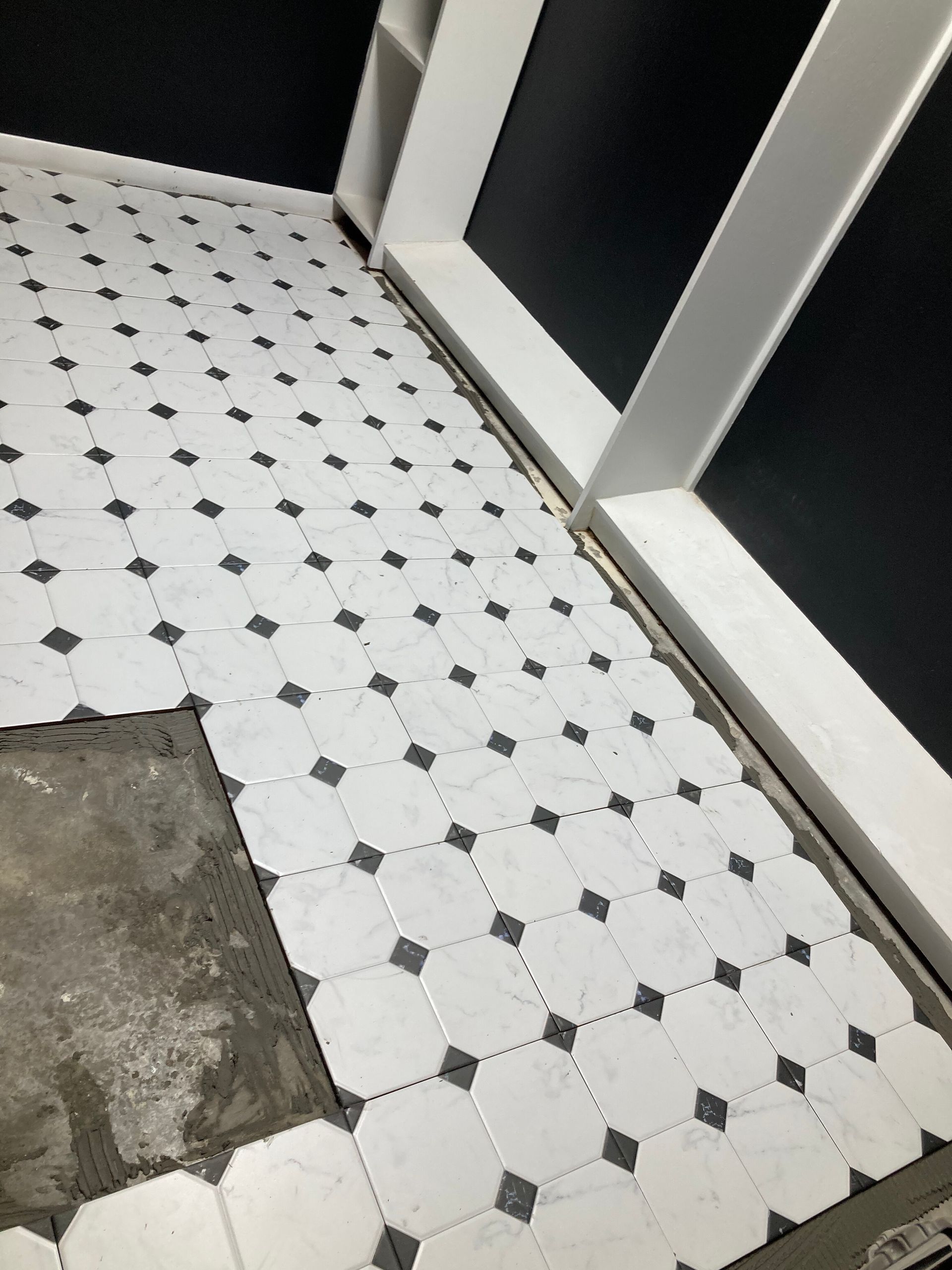 Black-and-white tiled floor with a square patch of exposed concrete near a wall and doorway