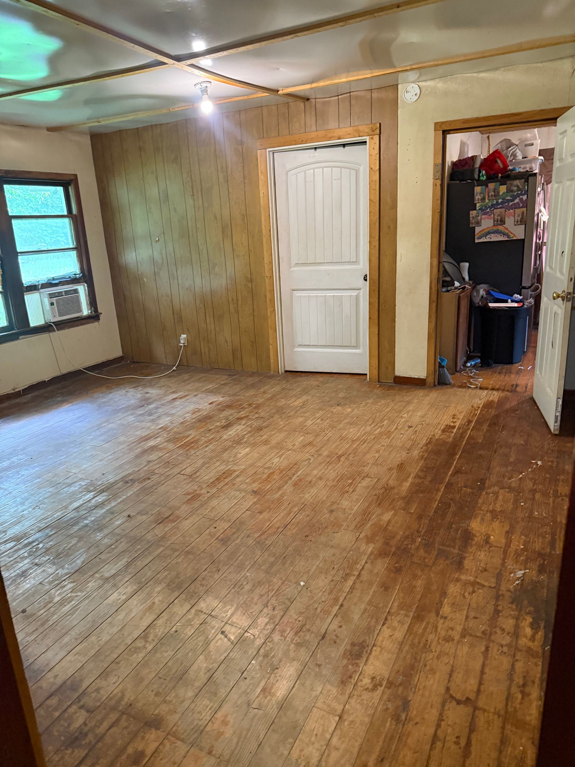 Empty worn room with wood floors, wood-paneled walls, a window, and two open doorways.