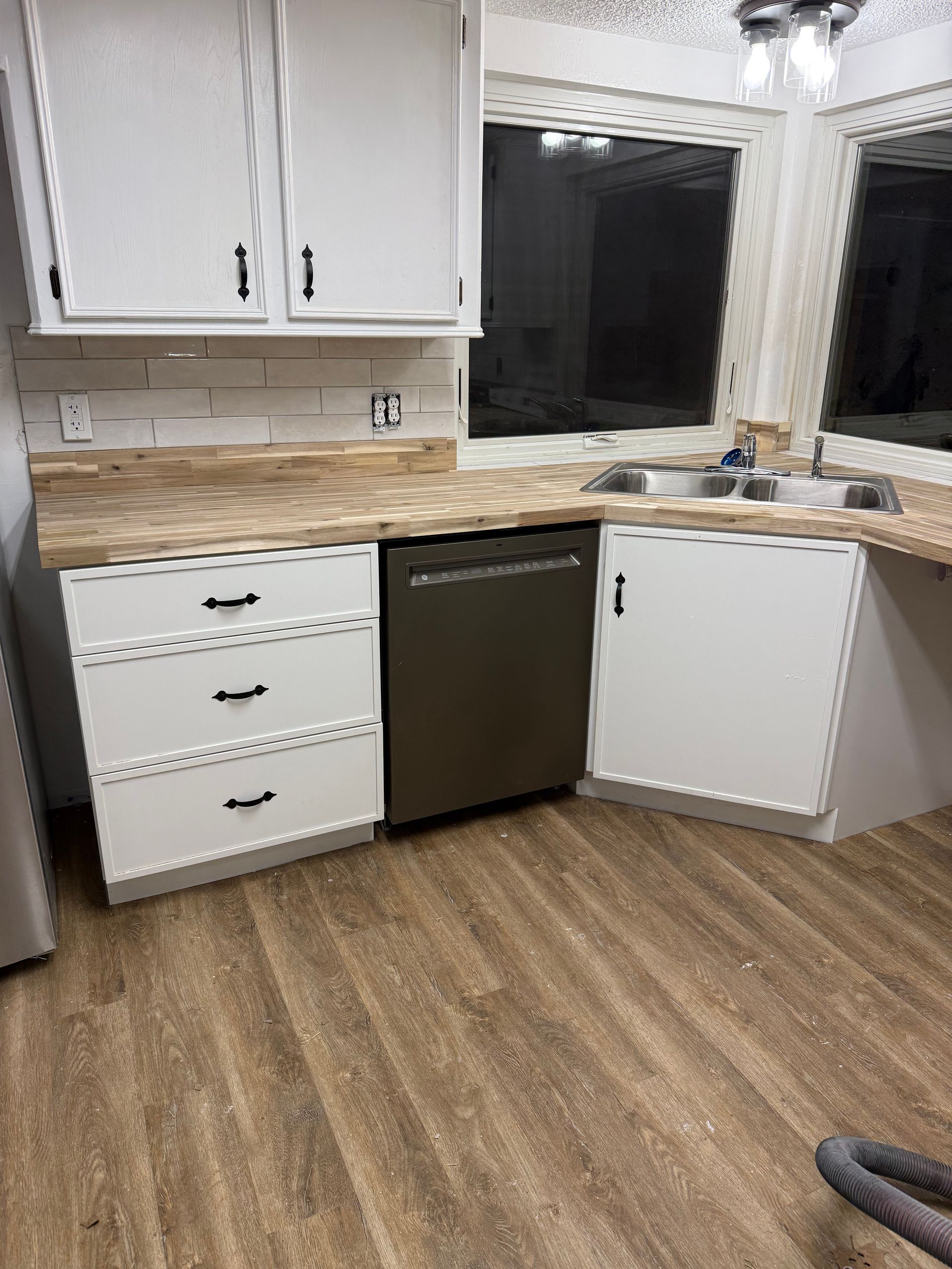 Kitchen corner with white cabinets, dark dishwasher, wood floor, and beige countertop under two windows