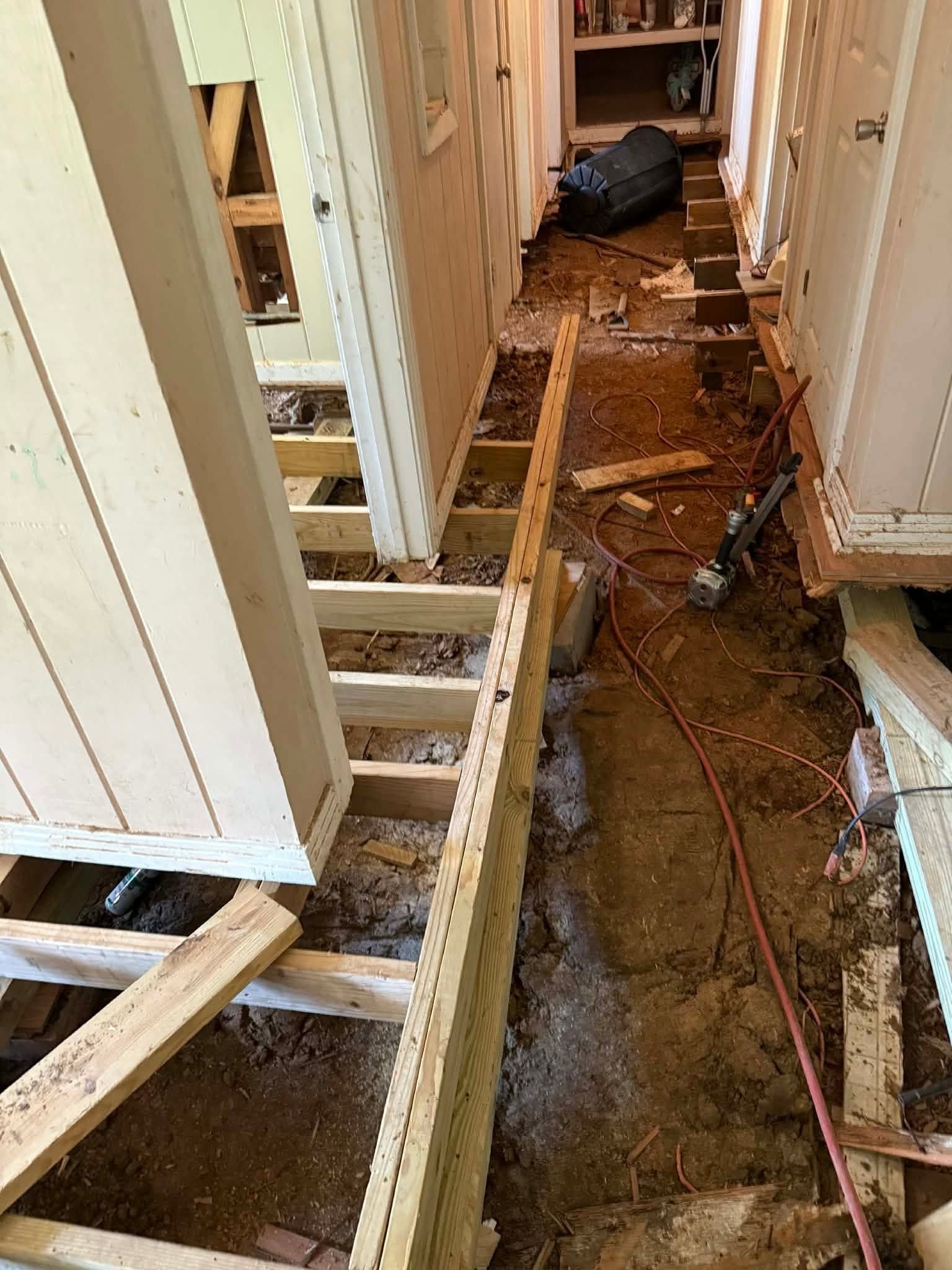Narrow hallway under home renovation with exposed floor joists, dirt, tools, and a red extension cord.