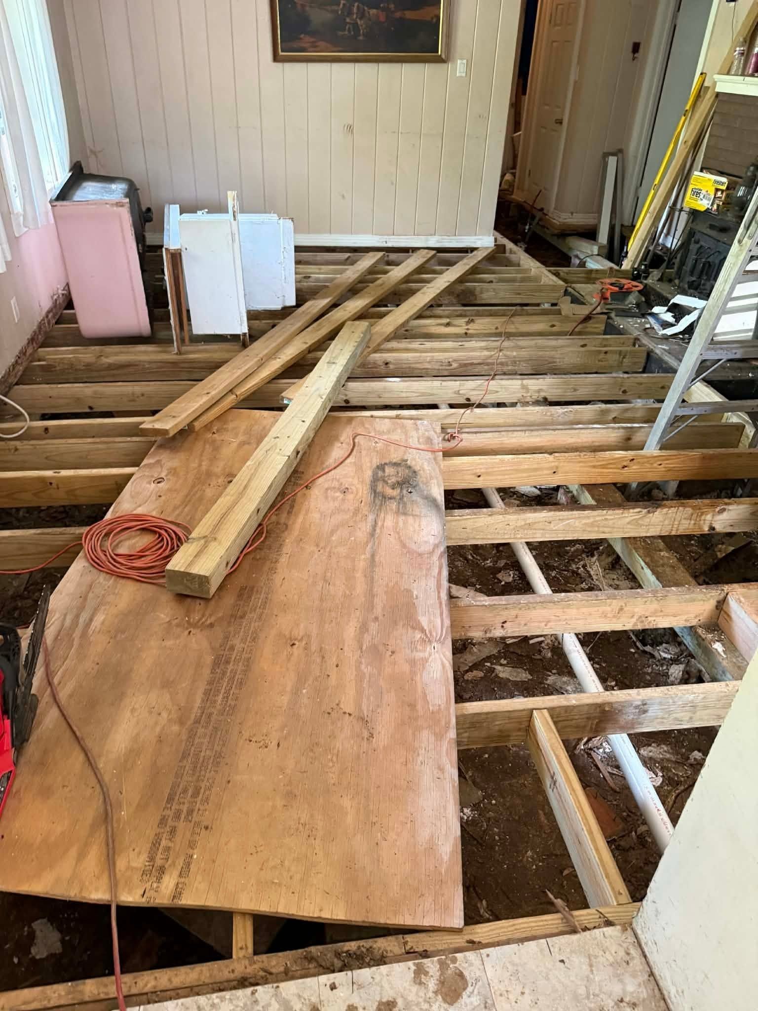 Renovation site with exposed floor joists, plywood sheets, and lumber laid across an unfinished room