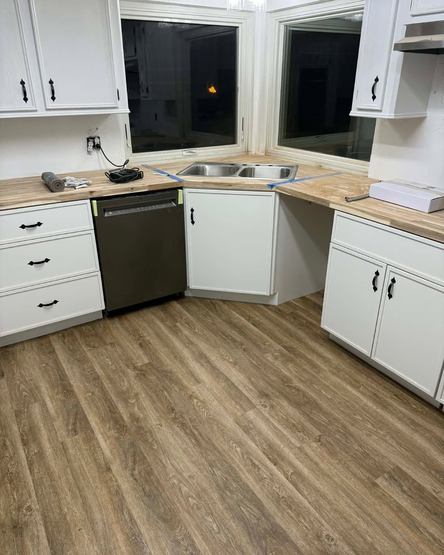 Modern kitchen with white cabinets, black dishwasher, corner sink, and wood-look flooring at night