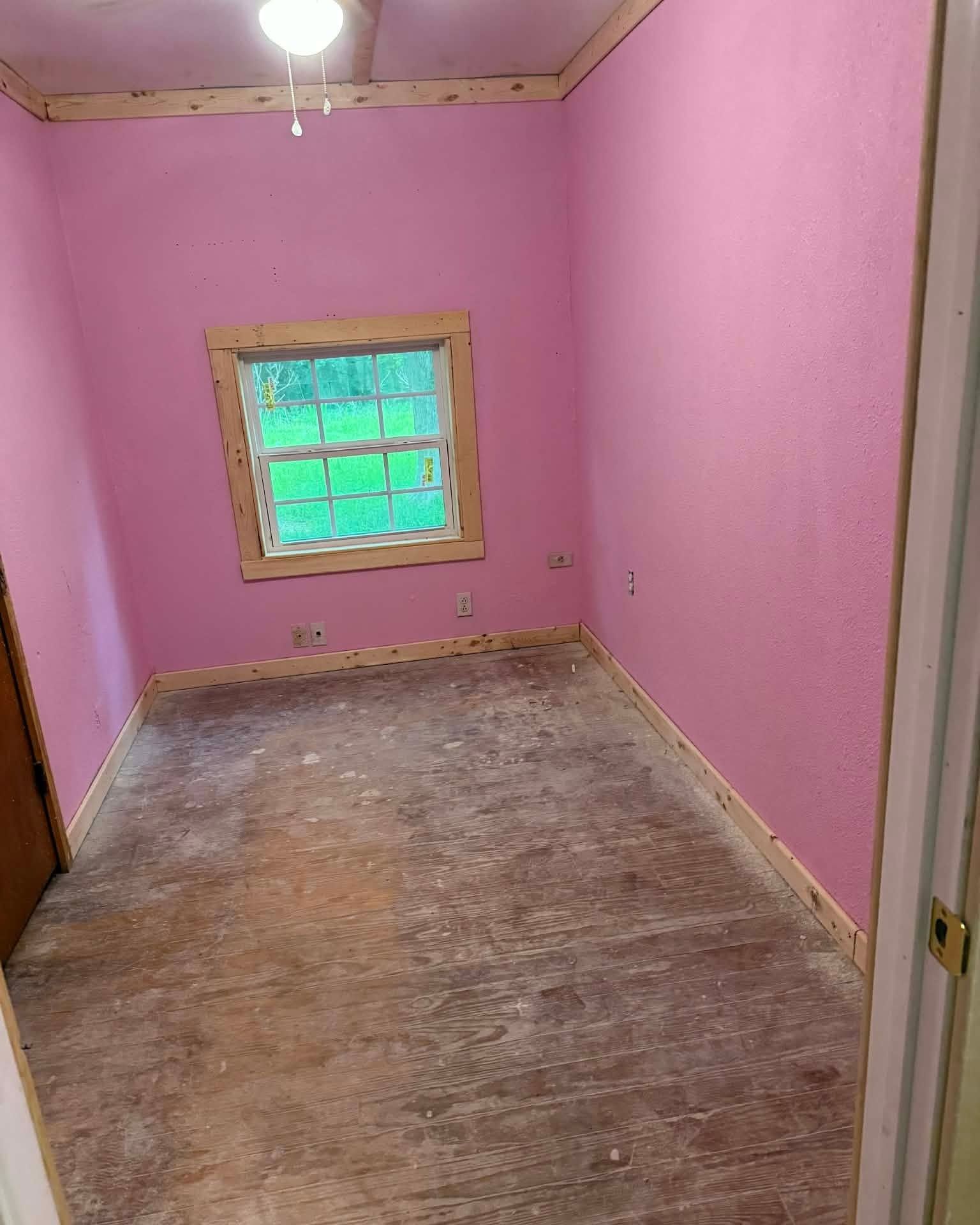 Empty pink room with worn floor and small green-paned window.