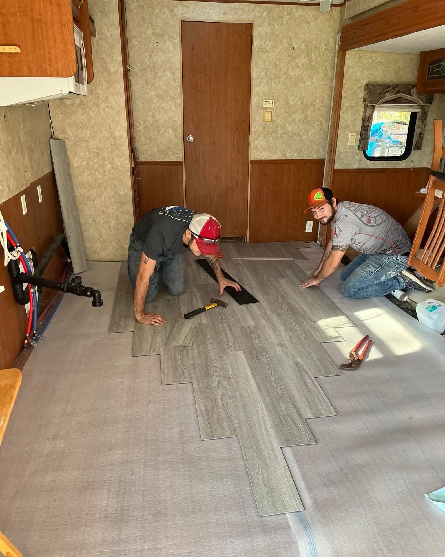 Two workers installing gray carpet tiles in a small room with wood-paneled walls.