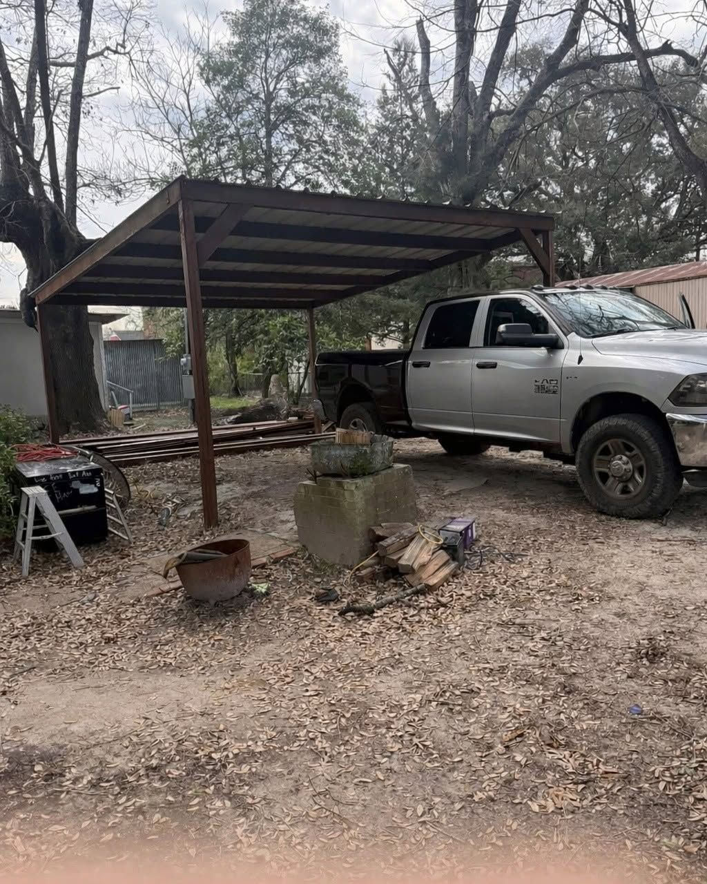 Silver pickup truck under a metal carport in a gravel yard, with trees and a fire pit nearby