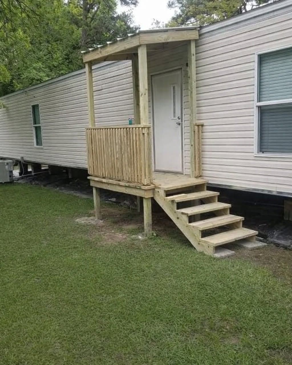 Small wooden porch with steps beside a beige mobile home on a grassy lot.