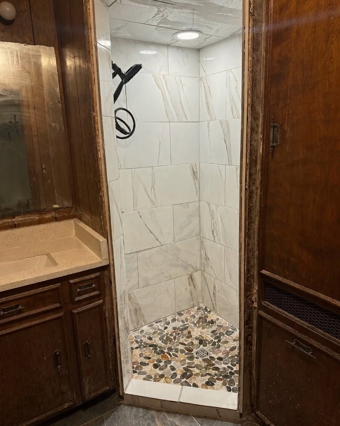 Open shower stall with white marble tile and pebble floor in a wood-paneled bathroom