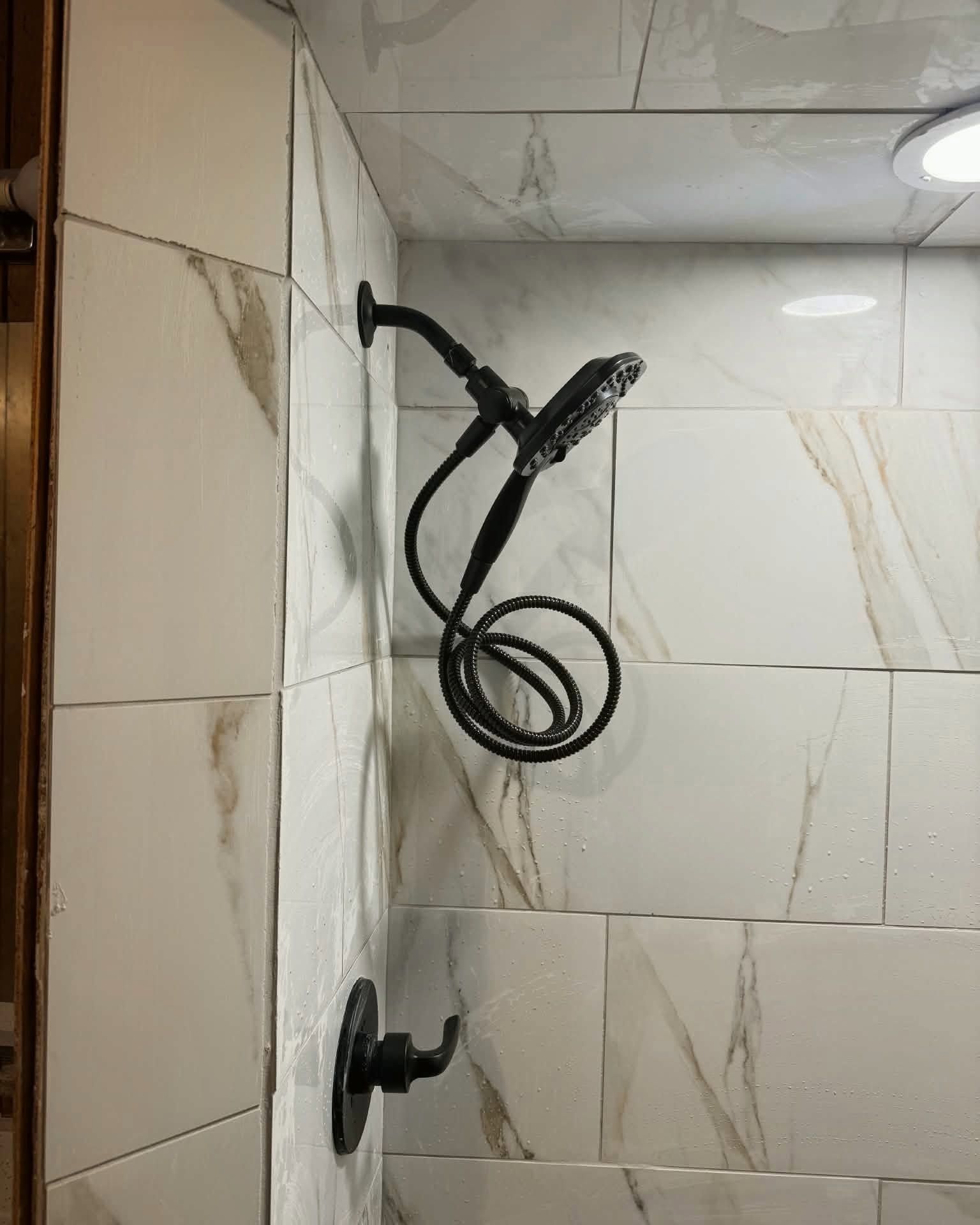 Bathroom shower corner with black handheld shower hose and wall-mounted fixture on white tiled walls.