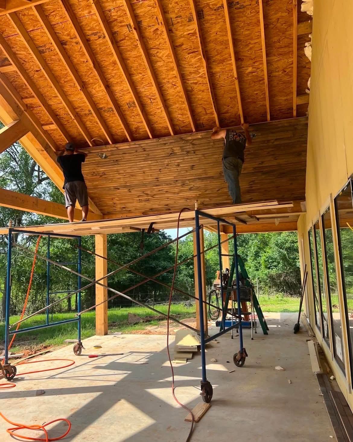 Two workers under a wooden roof, installing woven ceiling panels on scaffolding at a construction site.