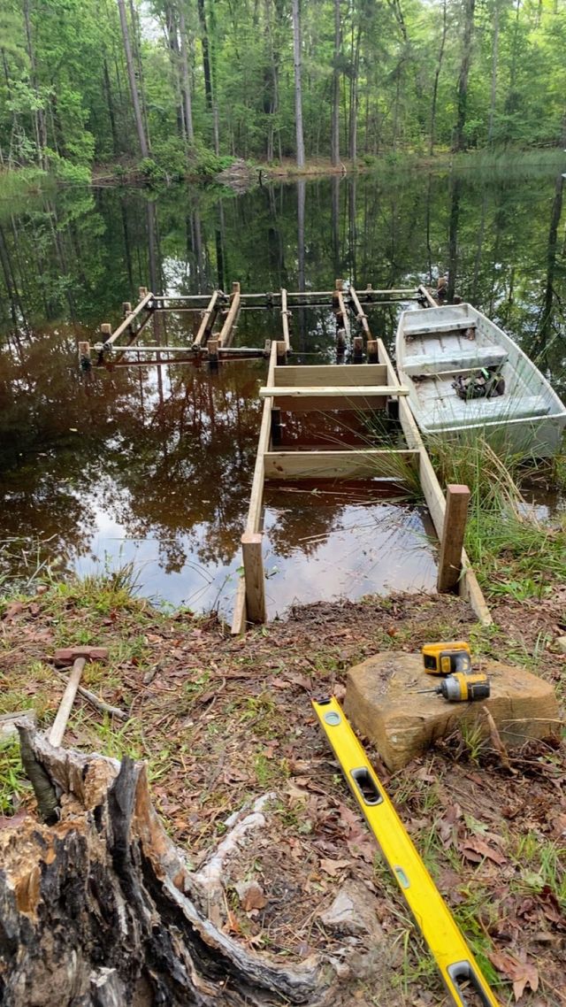 Damaged wooden dock extending into a wooded pond, with construction tools in the foreground