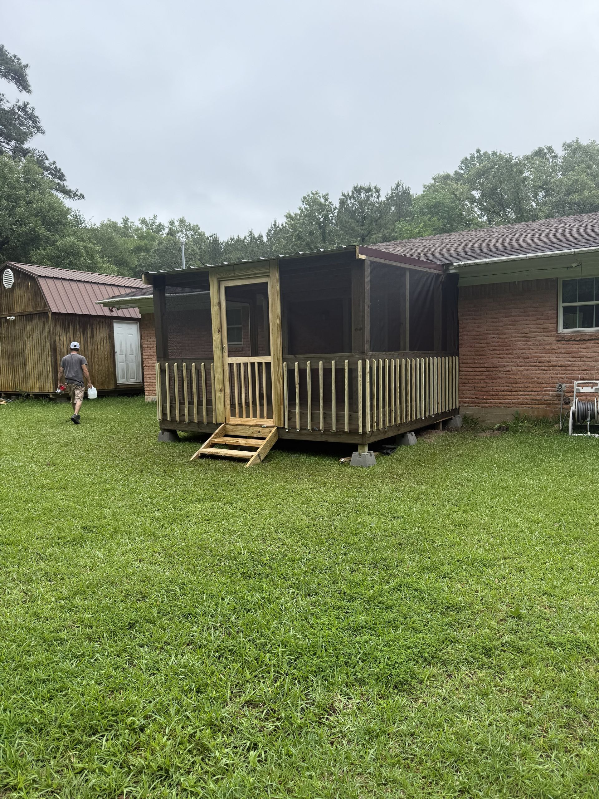Small wooden porch attached to a brick house in a grassy yard, with a person standing in the background.