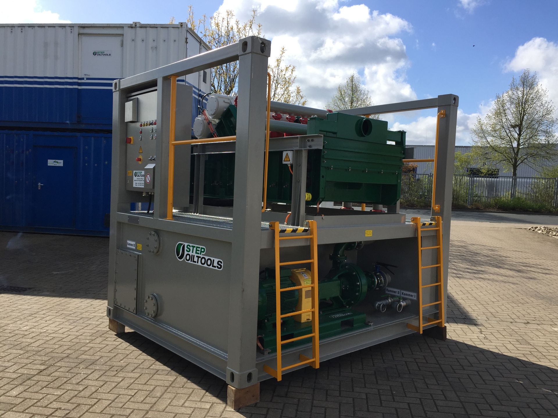 Industrial machine in a gray shipping container frame, with green equipment and yellow ladders outdoors