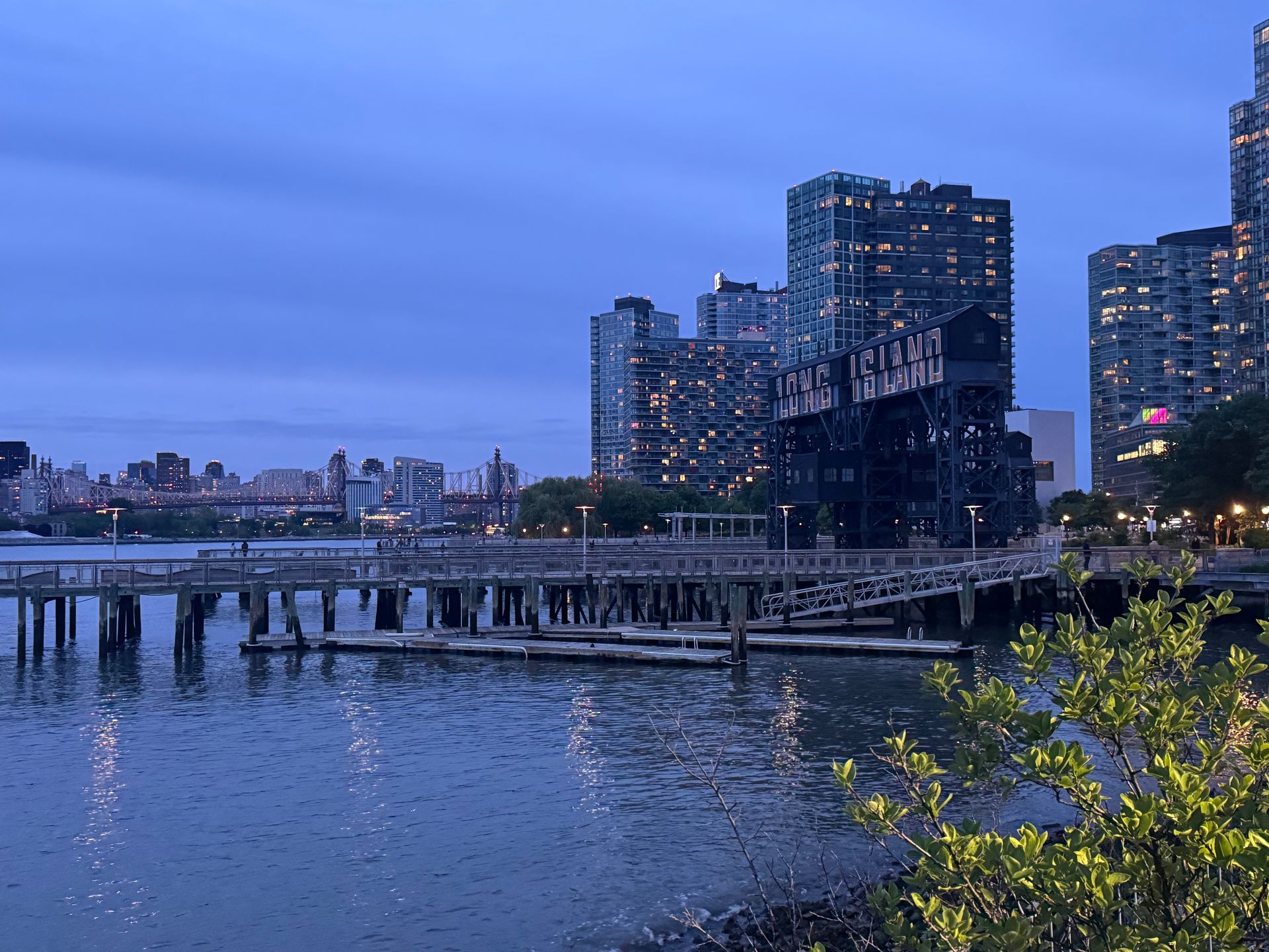 City waterfront at dusk with high-rise buildings, a lit bridge, and calm water reflecting the blue sky