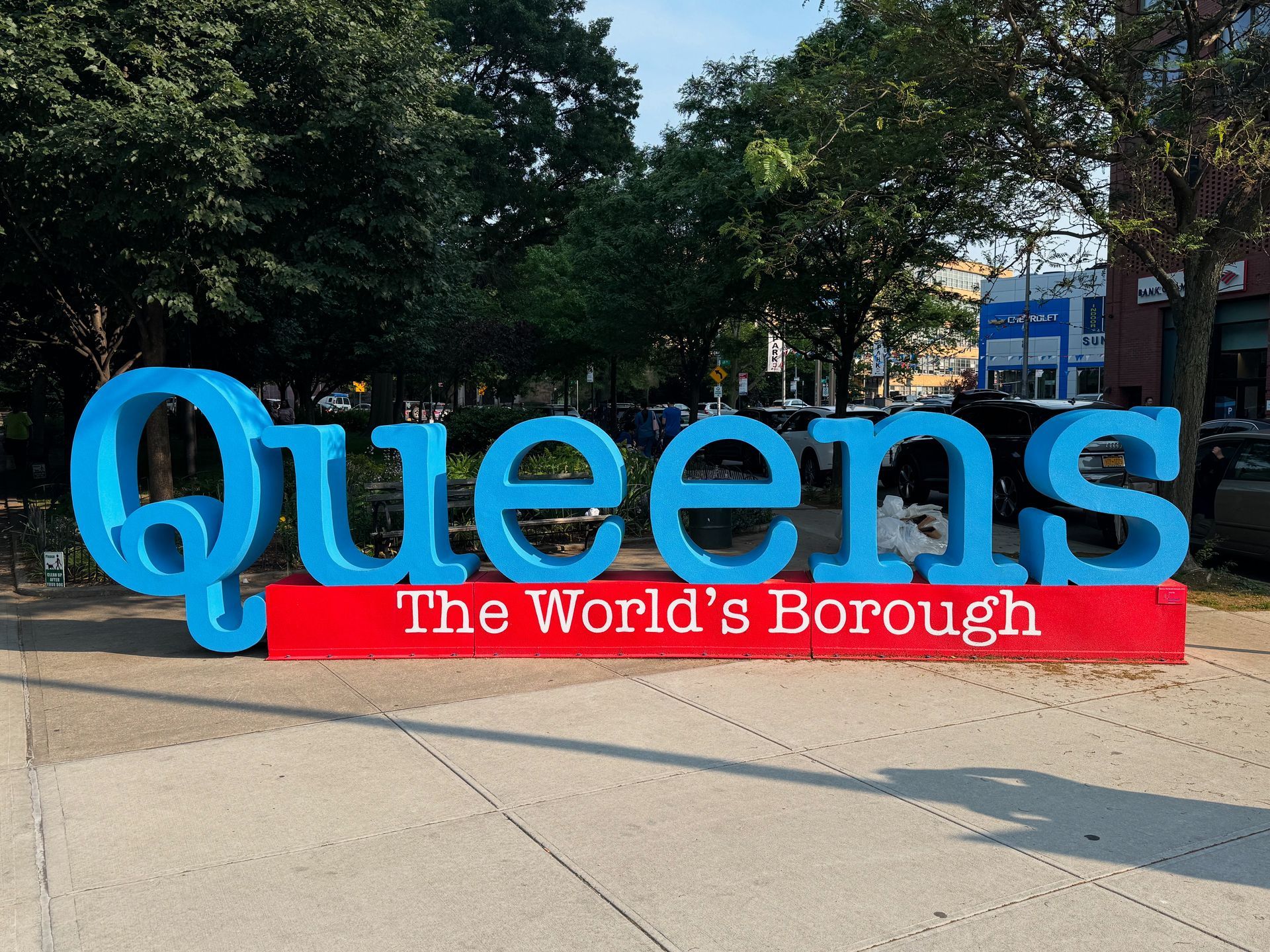 Blue “Queens” sign with red “The World’s Borough” banner in a park setting