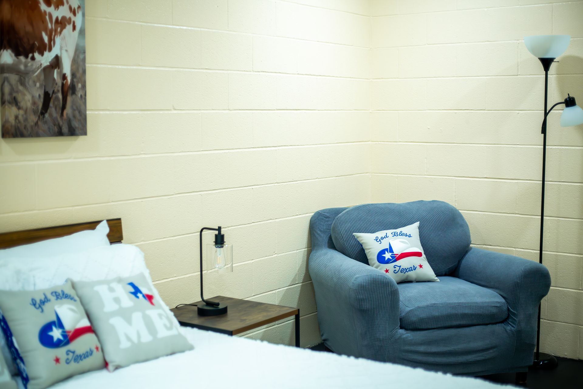 A bedroom with a bed , chair , lamp and a painting of a cow on the wall.