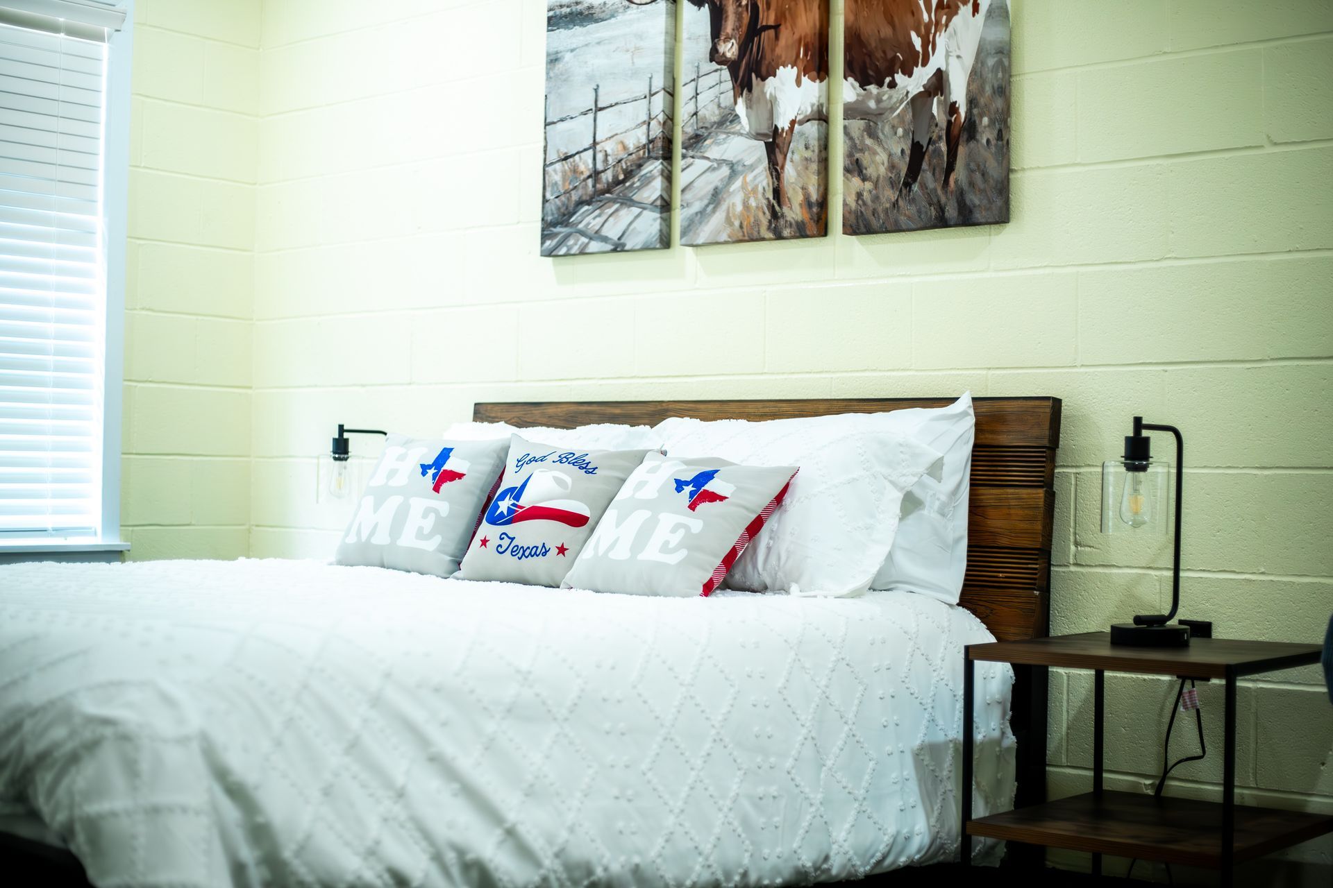 A bedroom with a bed , nightstand , lamp and paintings on the wall.