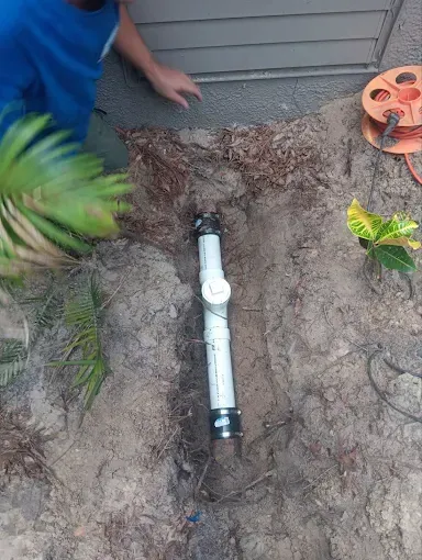 Excavated yard showing a white PVC pipe and joint, with a person kneeling beside it and a hose reel nearby