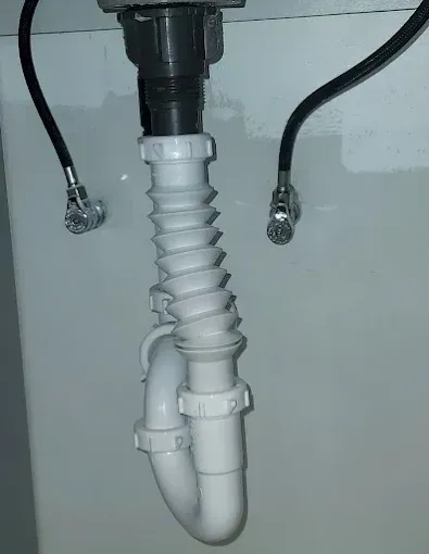 Under-sink white plastic drain trap with corrugated pipe and black supply lines on a wall