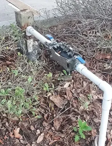 White PVC pipe with a small black valve assembly beside a concrete post in a leaf-covered yard