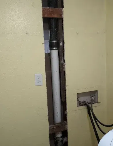 Open wall cavity with white pipe, electrical outlet, and cables in a yellow-painted room.