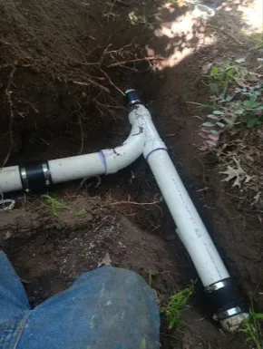 White PVC pipe junction in a dirt trench with black couplings and a blue tarp in the foreground