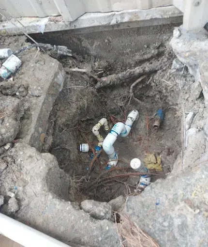 Broken underground pipe in a dug-out trench, with exposed plumbing and muddy soil around it.