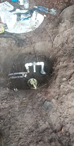 Excavated dirt pit with exposed black pipe and white fittings at the bottom.