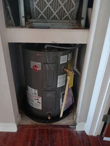 Water heater in a closet under a vent, with a metal tank and red hardwood floor.