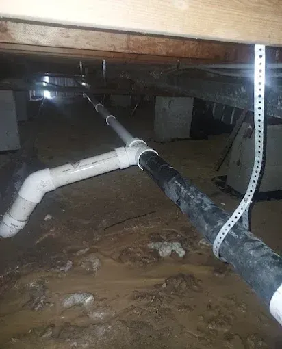 Flooded crawlspace with muddy water and PVC pipes under a low wooden floor.