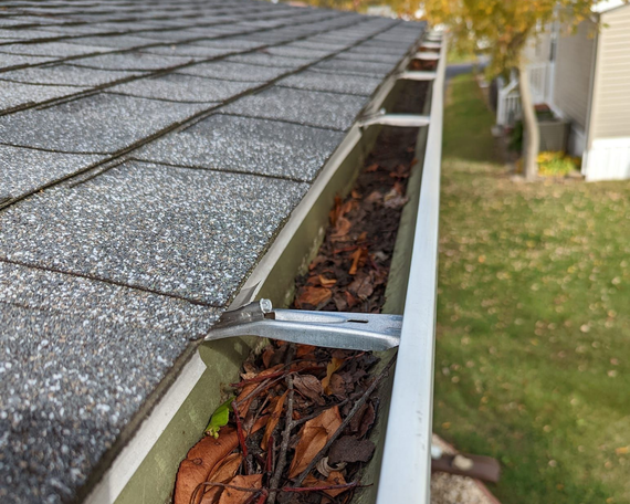 Gutter filled with leaves, attached to a shingled roof. Green grass and a house are in the background.