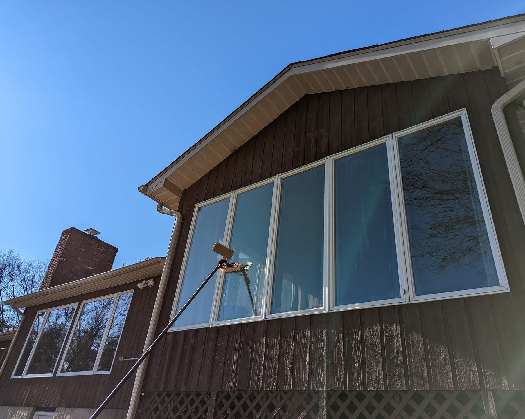 A person cleans large windows on a brown house exterior with a long-handled squeegee under a bright blue sky.