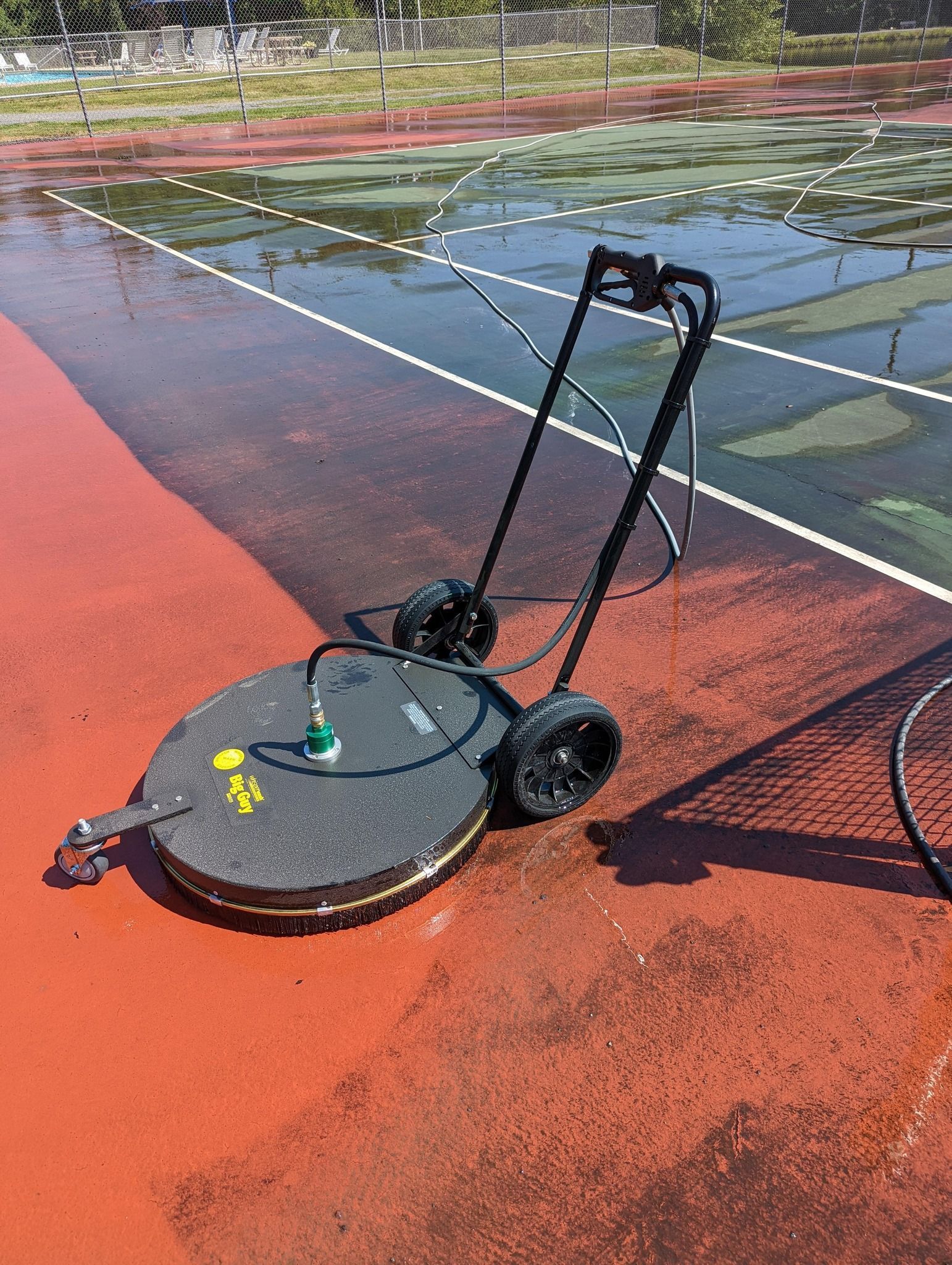 Tennis court being pressure washed with a circular surface cleaner. Red and green court surfaces, black machine.