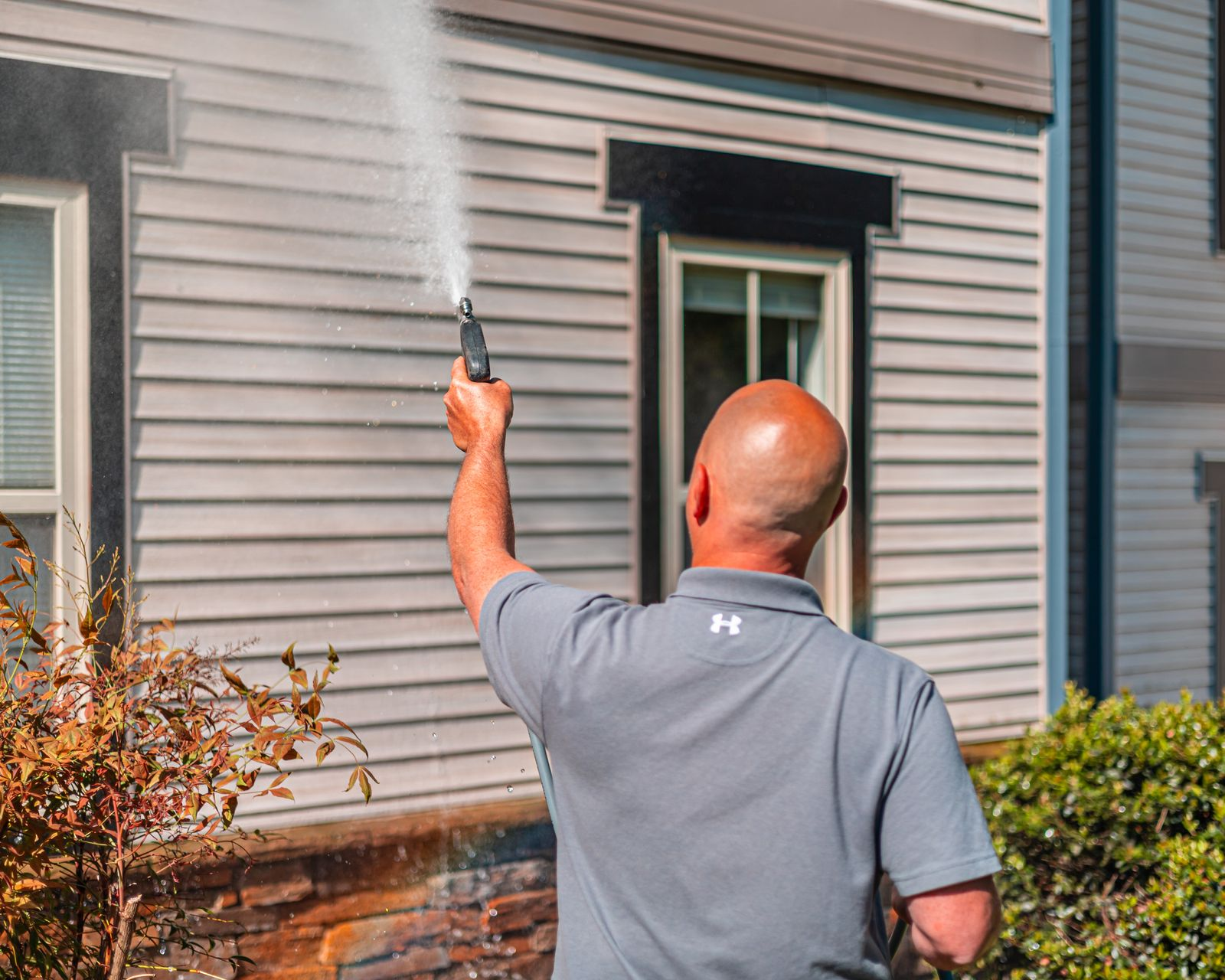 Man pressure washing a house's siding, aiming the spray upward. Focus on his bald head and back.