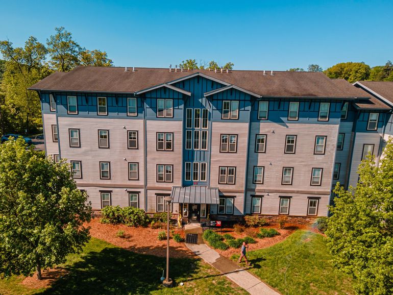 Multi-story apartment building with blue and gray siding, trees, and a walkway on a sunny day.