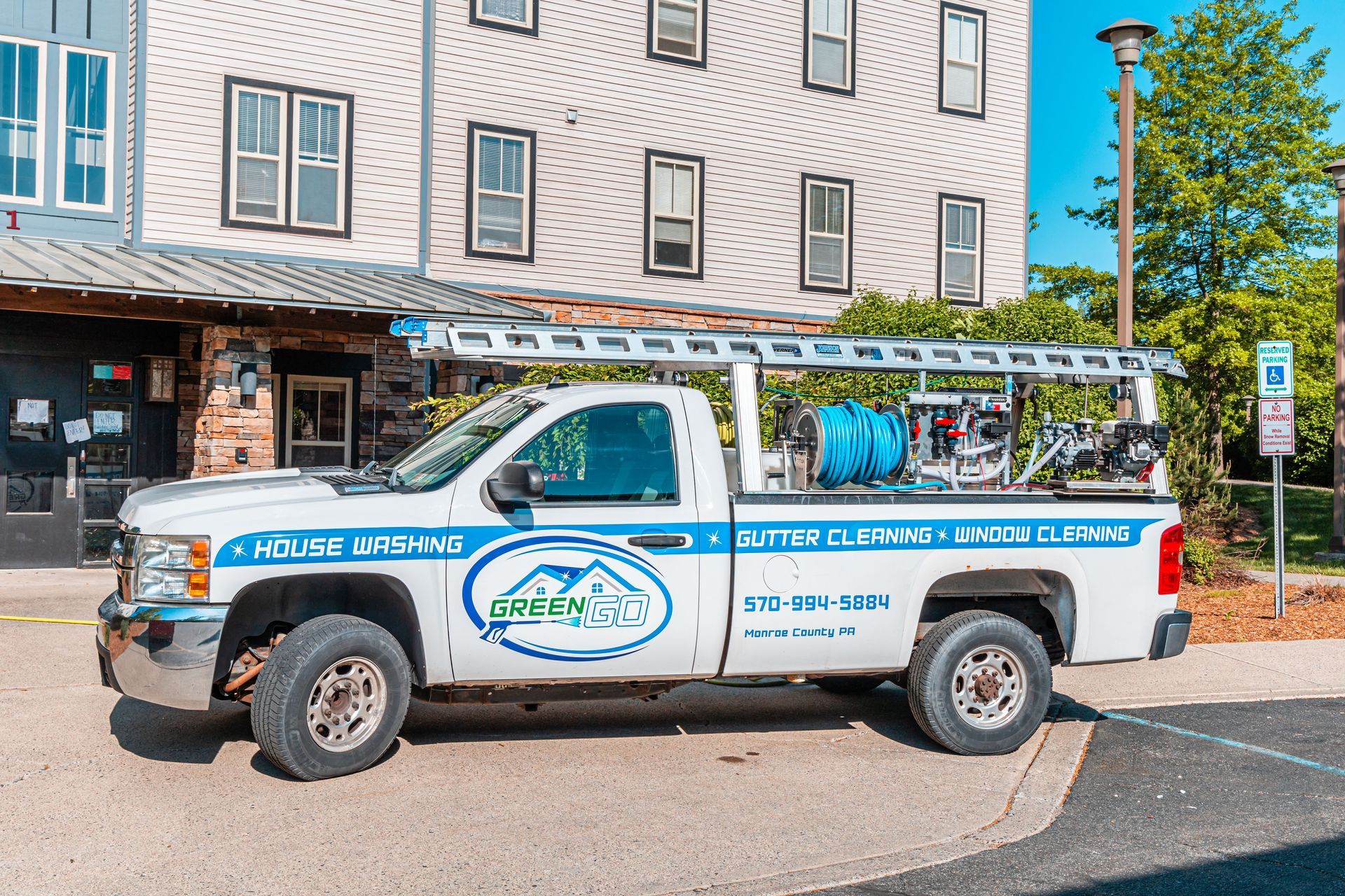 White truck with a ladder on top, parked in front of a building. The truck has a company logo on the side