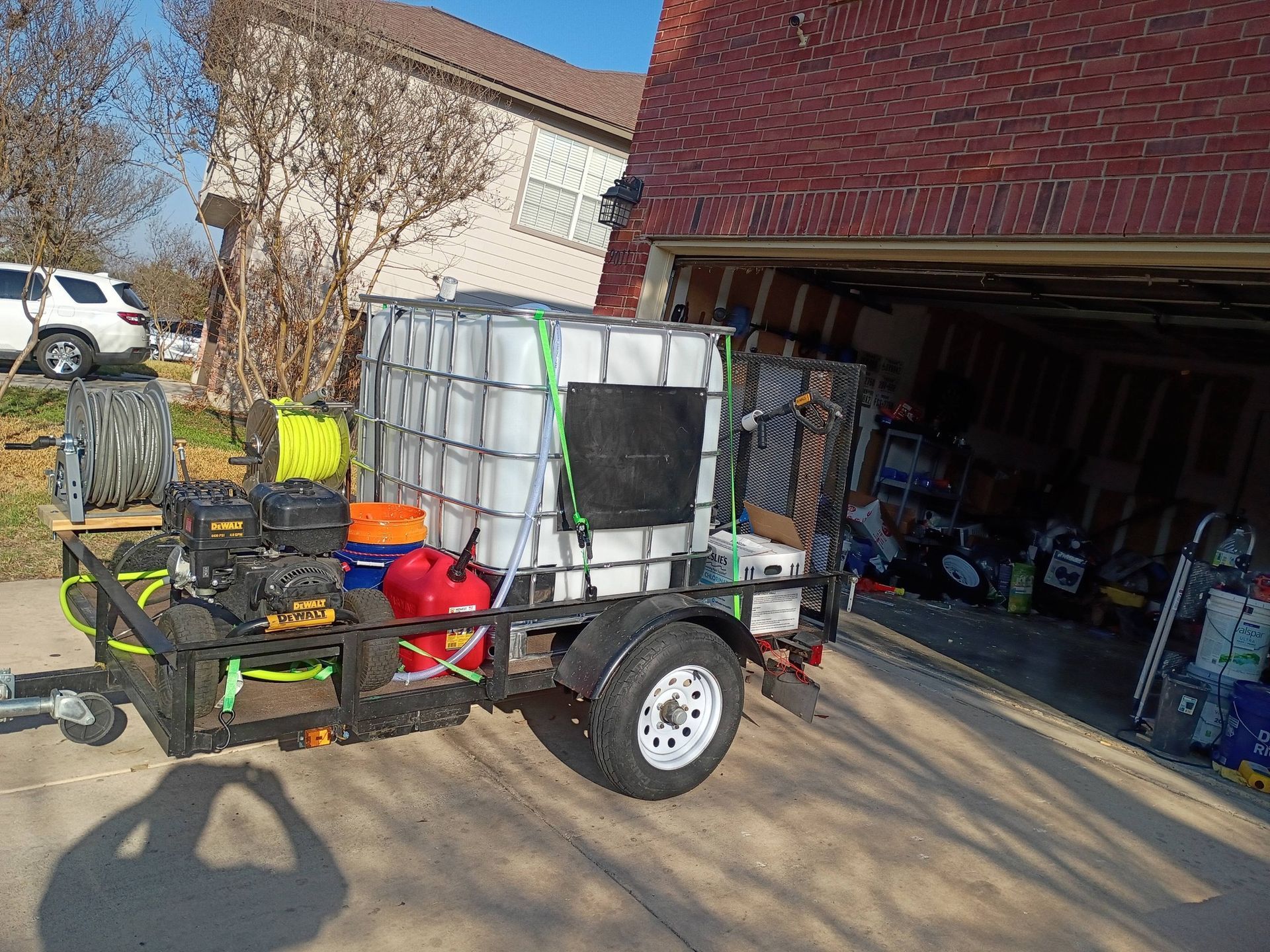 A utility trailer parked in a driveway, carrying a large white water tank, a power washer, and various equipment.
