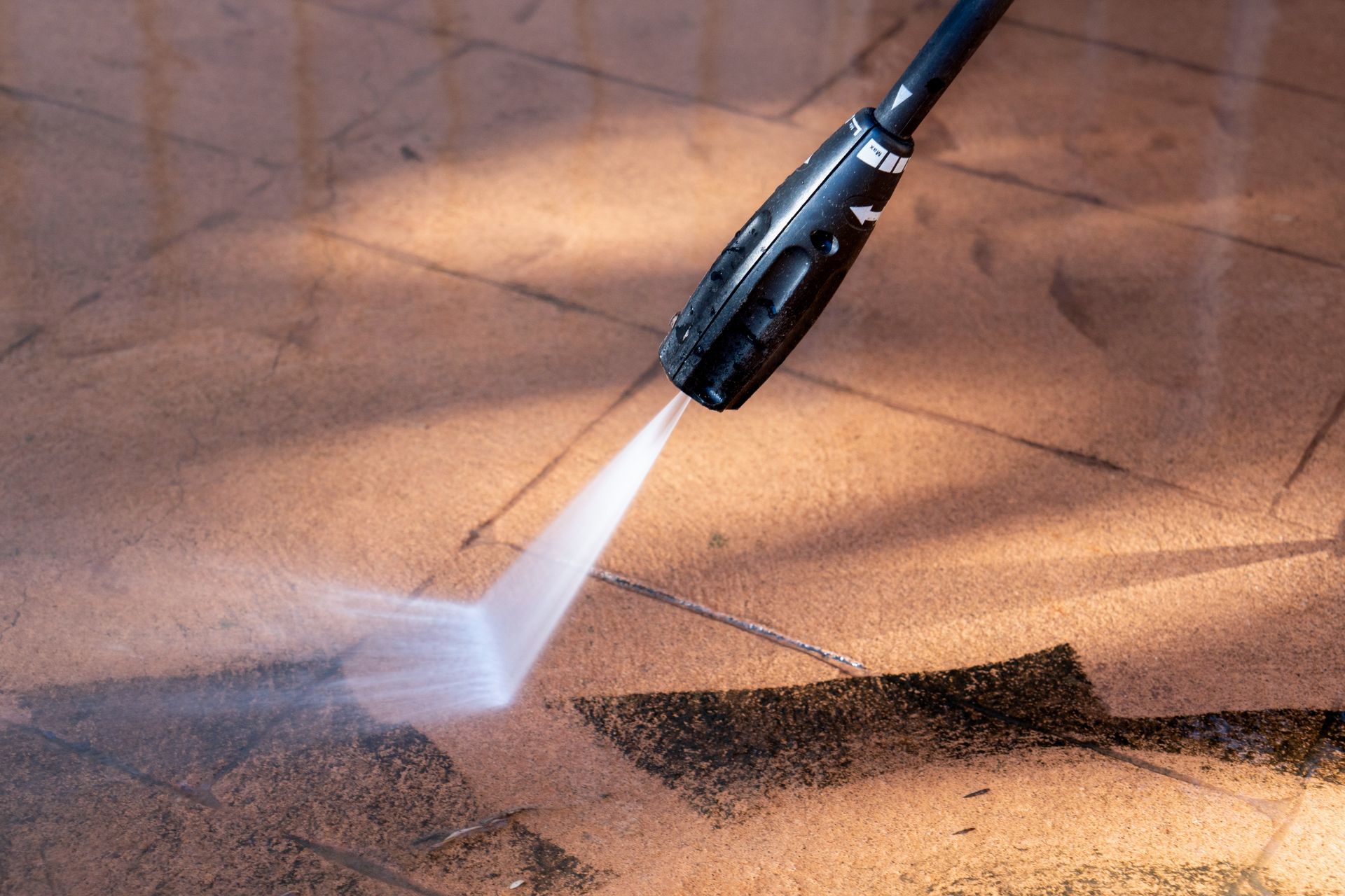A high-pressure washer nozzle sprays a fan of water onto a stained tiled floor, cleaning a dark patch of grime.