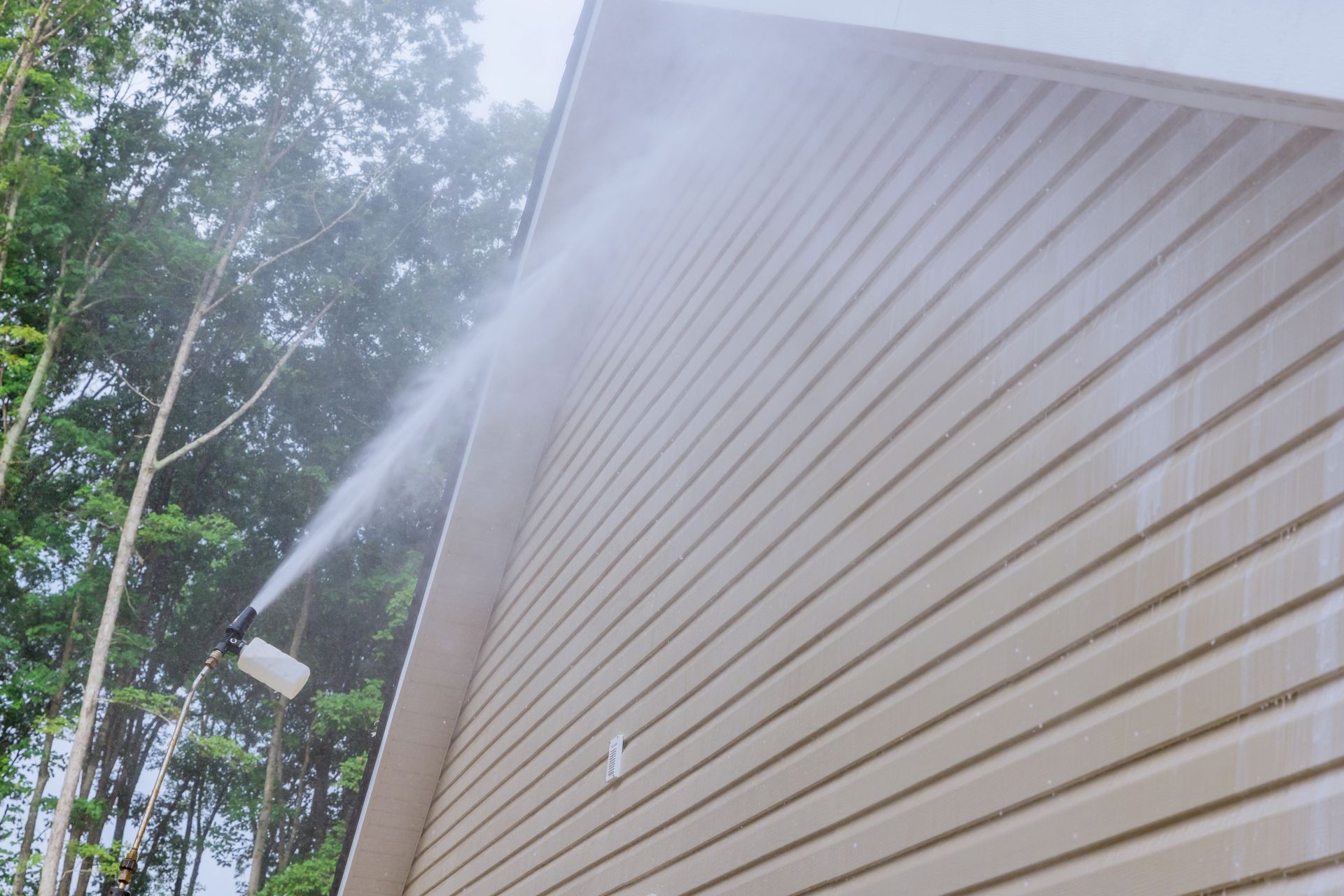 A pressure washer sprays cleaning solution onto the beige vinyl siding of a house exterior near trees.