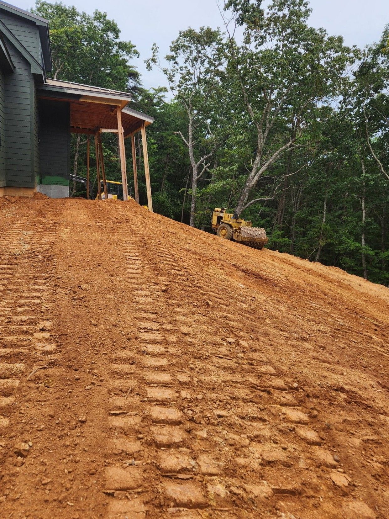 Freshly graded dirt driveway beside a house, with a small bulldozer and wooded hillside.