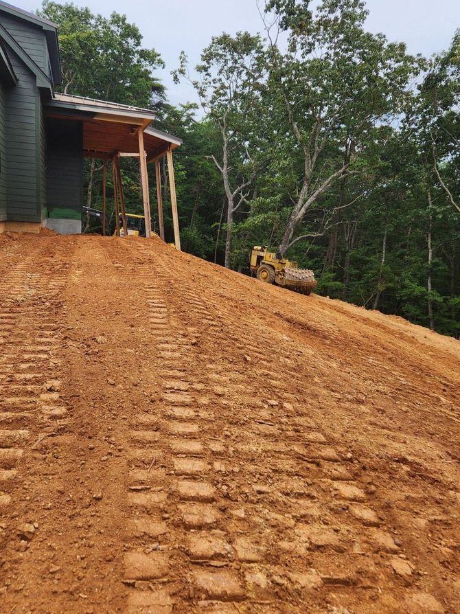 Freshly graded dirt slope beside a house, with tire tracks and a bulldozer in the distance.