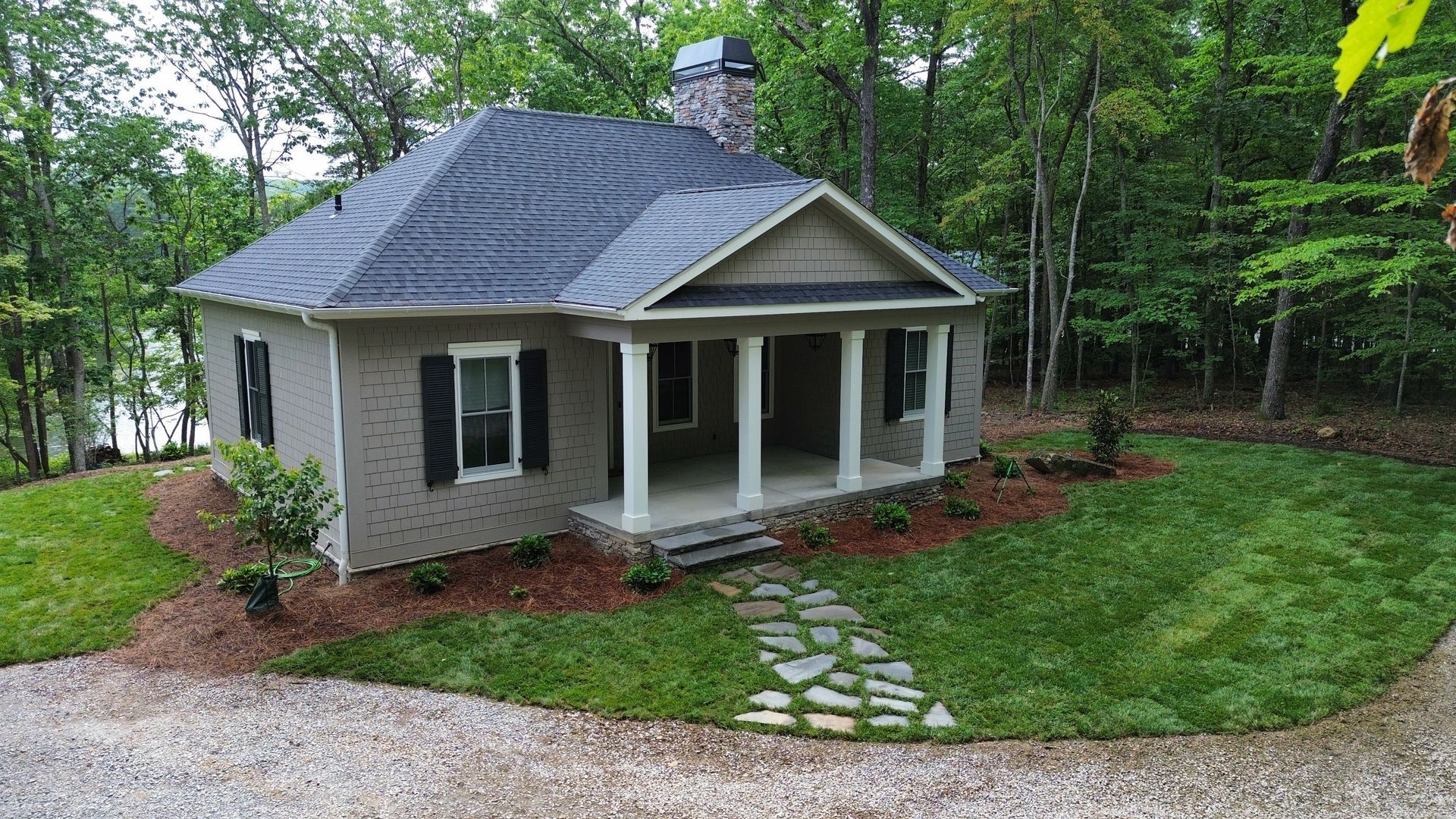 Small stone house with front porch, gravel driveway, and green yard surrounded by trees