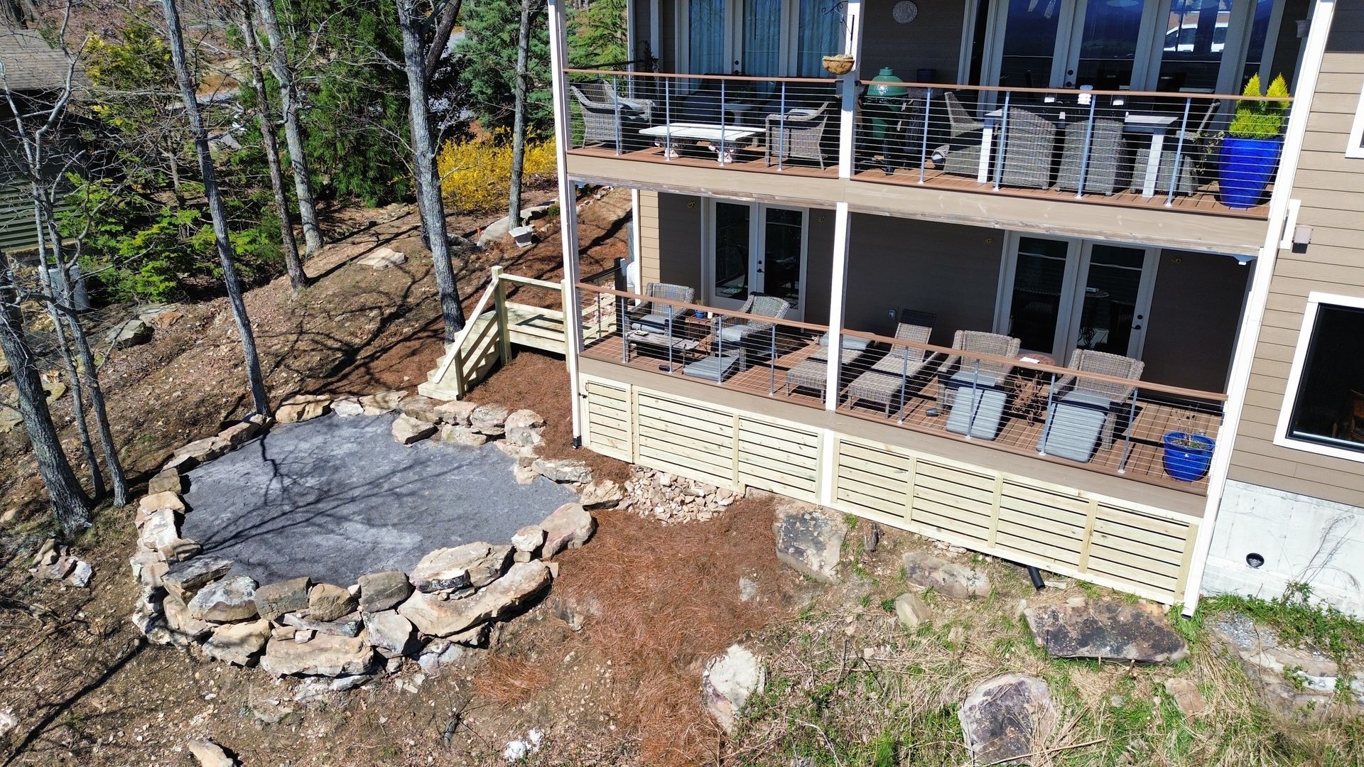 Outdoor deck and patio with metal railings, a stone fire pit, and chairs beside a wooded hillside.