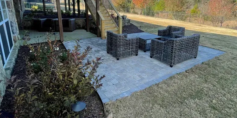 Backyard patio with wicker seating around a fire pit on a paved area, beside a garden bed