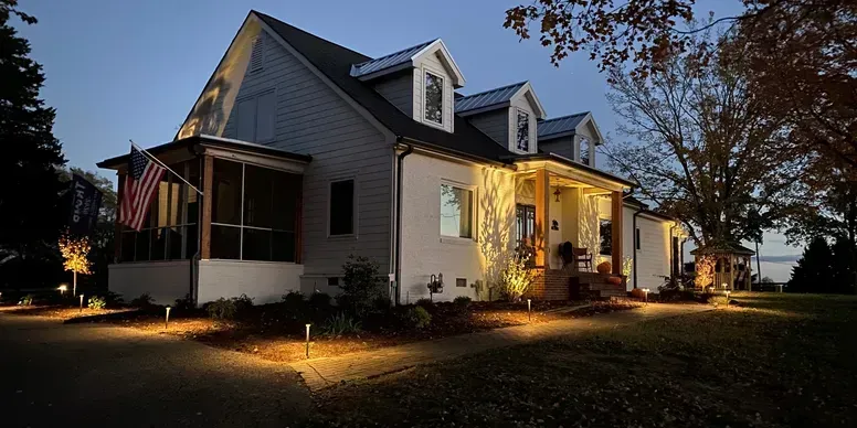 Lit white house glowing at dusk with warm porch lights and autumn trees nearby
