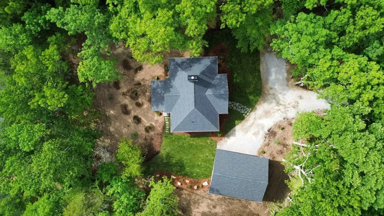 Aerial view of a house with a dark roof surrounded by dense green trees and a dirt driveway