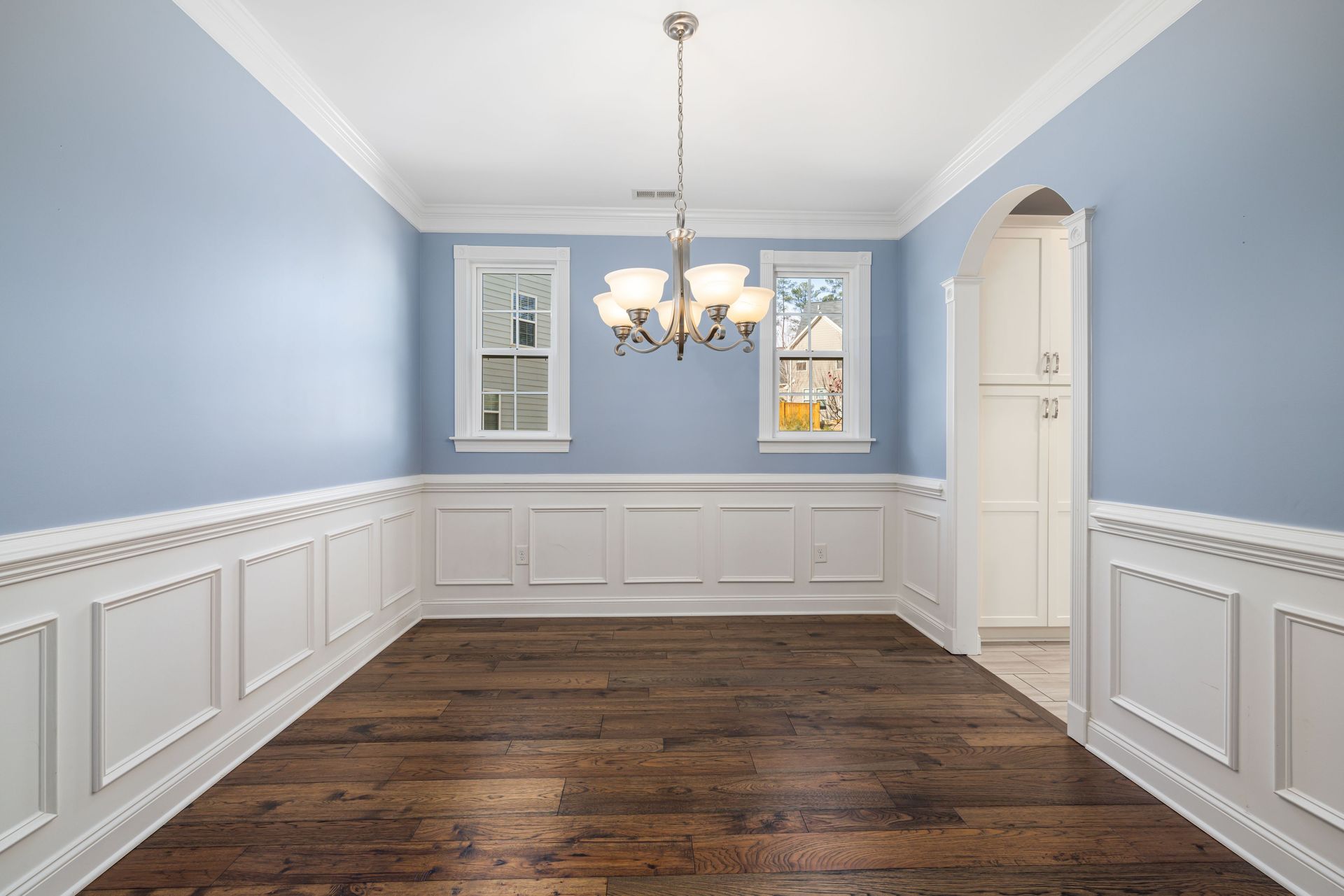 Empty dining room with light blue walls, white wainscoting, dark wood floor, and a chandelier
