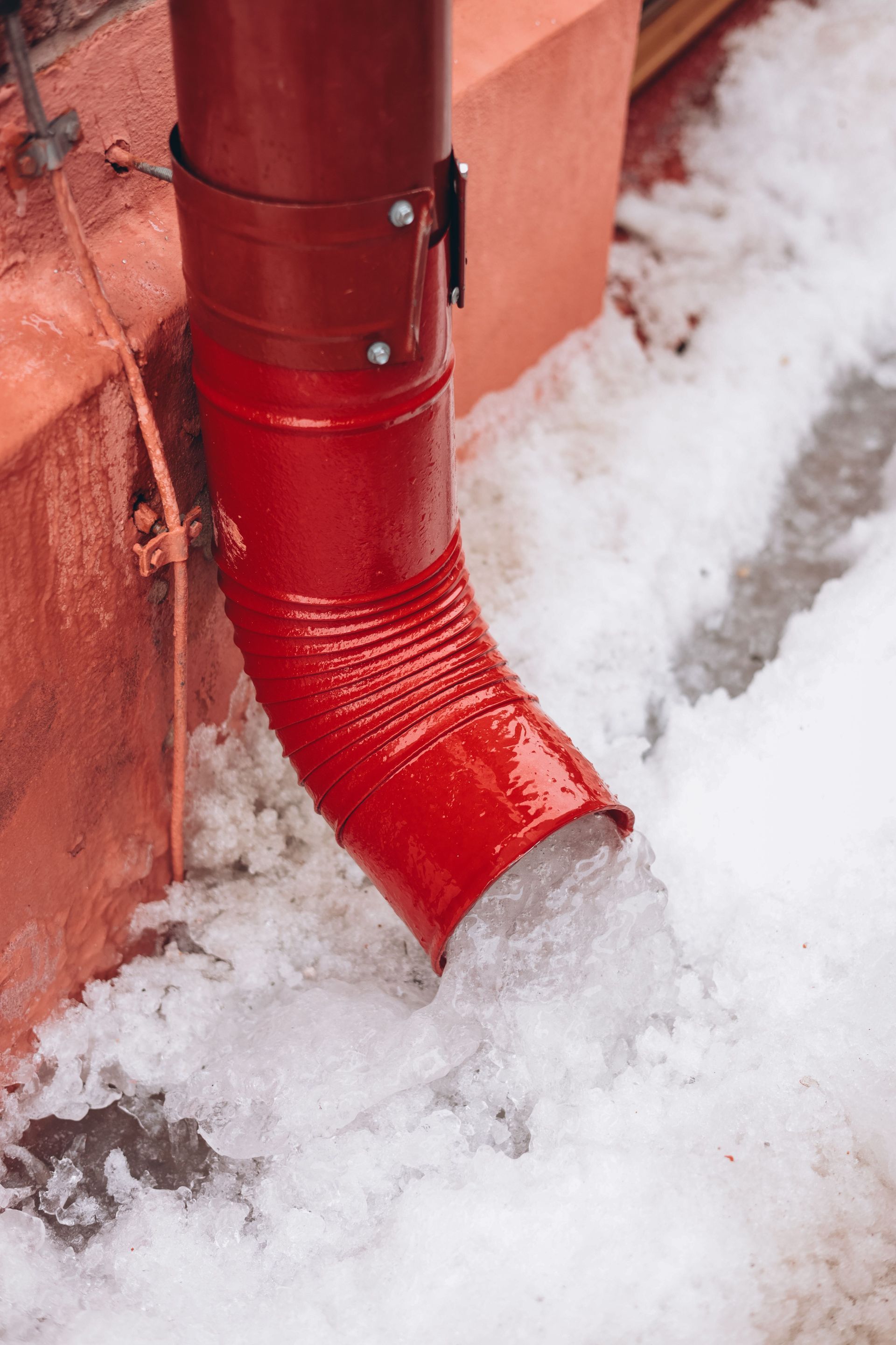 Red downspout draining into snow beside an orange wall