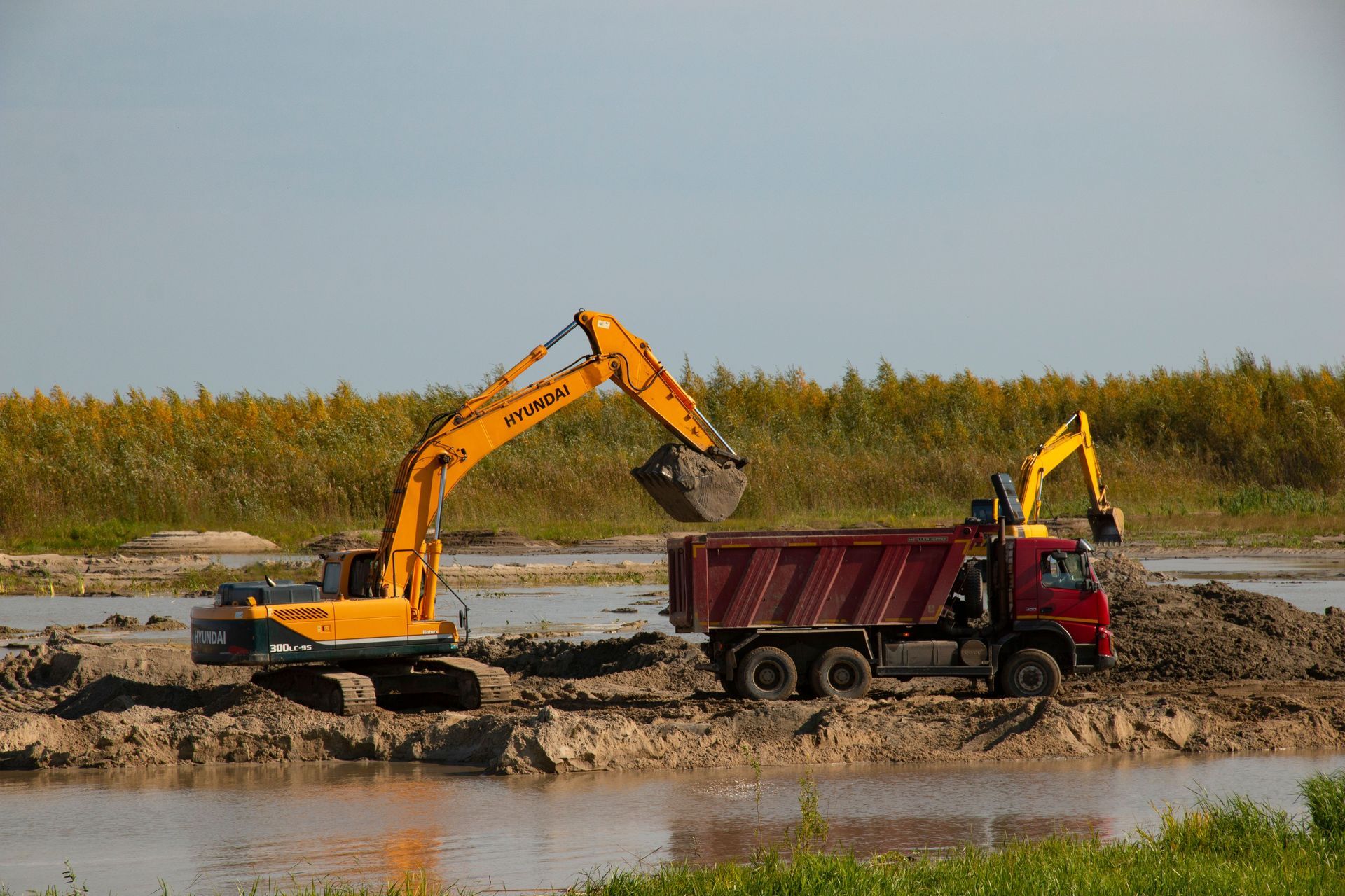 Yellow excavator loading dirt into a red dump truck at a muddy construction site by water.